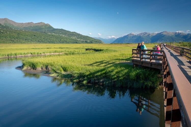 A wooden boardwalk with railings stretches across a marsh where visitors stop to take in the view. Still blue water reflects tall green grasses and the surrounding mountains, glowing in warm sunlight beneath a clear blue sky.