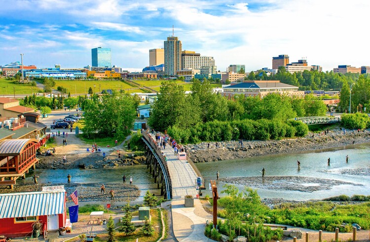 Summer view of Ship Creek in downtown Anchorage, Alaska, where anglers in waders fish the shallow water and visitors crowd a wooden footbridge. A red fishing rentals shop flying U.S. and Alaska flags sits in the foreground, with green trees, the historic Alaska Railroad depot, and the downtown hotel and office skyline rising under a bright blue sky.