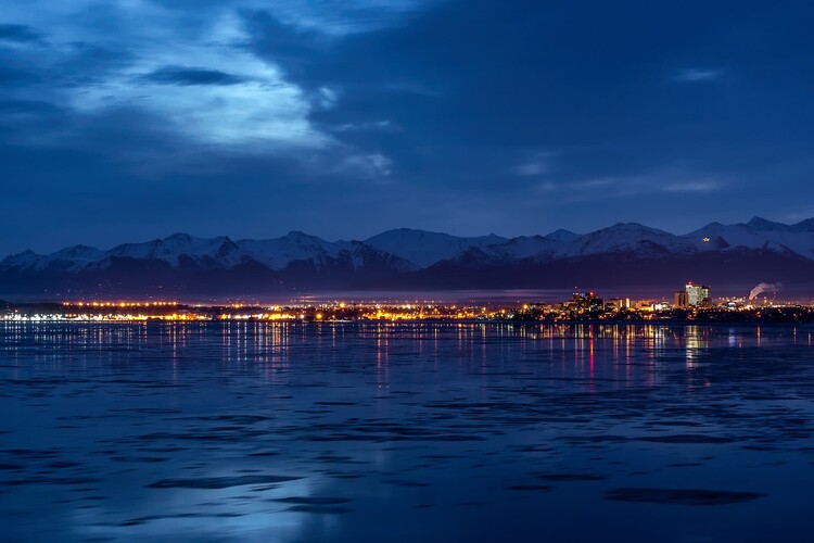 Nighttime winter view of downtown Anchorage, Alaska, seen across the icy waters of Cook Inlet. City lights stretch along the shoreline and reflect in shimmering streaks on the water, while the snow-capped Chugach Mountains glow softly beneath a deep blue sky streaked with clouds.