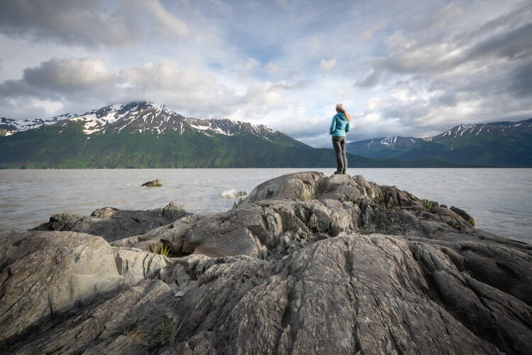 A woman in a teal jacket and gray pants stands atop weathered gray rocks at the water