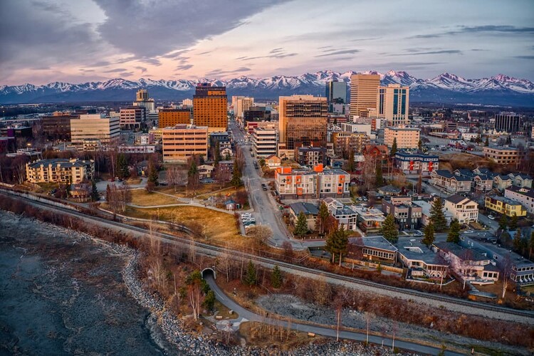 downtown Anchorage hotel An aerial skyline view of the city buildings and a prominent downtown Anchorage hotel with the snow-capped Chugach Mountains in the background at sunset.